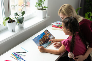 Happy teacher and student girl sitting at working desk in classroom. Cheerful young mother and child sitting at table and smiling. Little kid and her private language tutor