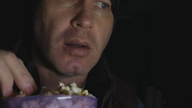 emotional Man watches a scary and interesting movie on TV in the dark and eats popcorn from a plastic bucket.