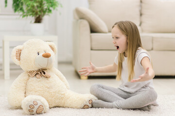 Photo of little blond girl talking to her white teddy bear.