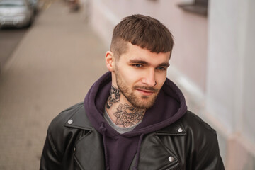 Fototapeta premium Street portrait of a handsome stylish hipster guy with a hair and beard with a tattoo on his neck in fashionable clothes with a jacket and hoodie walking in the city