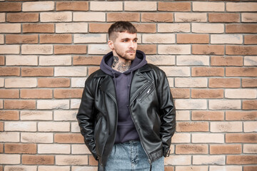 Fashionable hipster man with a tattoo on his neck in stylish casual clothes with a jacket and hoodie standing near a brick wall on the street