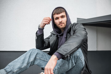 Stylish handsome young hipster man in fashionable clothes with hoodies and leather jacket and jeans sitting near the building on the street