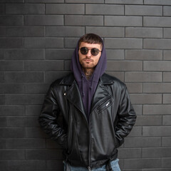 Urban stylish portrait of a handsome hipster man in fashionable clothing with a hoodie and leather jacket stands near a black brick wall in the city