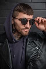 Obraz premium Fashion street male portrait of a handsome business stylish man in fashionable black sunglasses with a hood in a trendy black leather jacket and hoodie on a dark brick background on the street