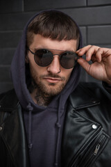 Fashionable hipster man model with hairstyle in stylish urban outfit with jacket and hoodie wears a fashion sunglasses near a brick black wall