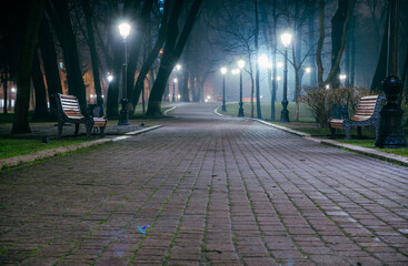 The alley of a night early winter park in a light fog. Footpath in a fabulous late autumn city park at night with benches and latterns. Beautiful cold evening in Mariinsky Park. Kyiv, Ukraine.