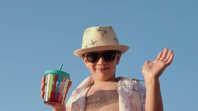happy boy in hat and sunglasses drinking juice on the beach