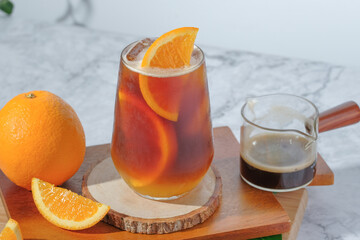 cold Iced black coffee mixed with orange juice and orange slices in glass on wooden plate in white background with nature leaves silhouette shadow. two tones layer fresh summer drink in the morning