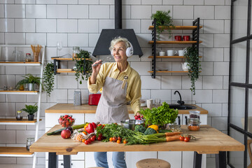 Senior attractive woman listening to music and singing in her headphones in the kitchen. Active lifestyle of mature people, enjoying life, having fun. Using whisk as microphone while cooking food