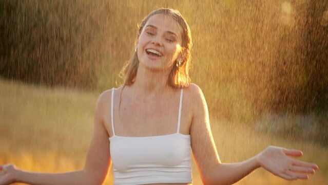 Young happy blonde woman wearing white top is feeling free and smiling under the rain. Stunning golden sun rays shine on playful young woman enjoying a summer rain