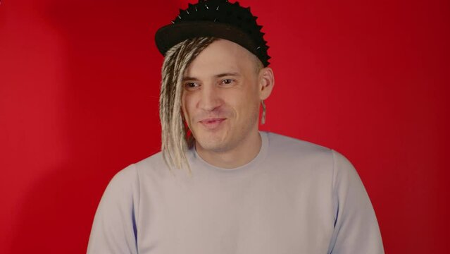 Young handsome man with dreadlocks in black cap talking, looking away, smiling, laughing on red background. Cheerful guy with happy facial expression discussing something in studio.