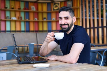 Portrait of happy positive Arab Muslim ethnic guy, young bearded man freelancer with black beard is drinking coffee sitting in cafe or restaurant working on laptop computer, smiling looking at camera 