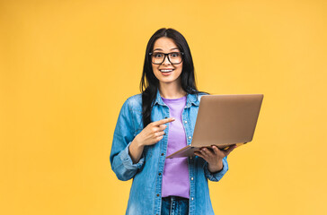 Portrait of happy young beautiful surprised woman with glasses standing with laptop isolated on yellow background. Space for text. - 514082924
