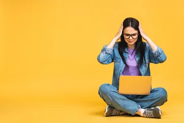 Fotobehang Business concept. Portrait of sad tired depressed woman in casual sitting on floor in lotus pose and holding laptop isolated over yellow background. © denis_vermenko