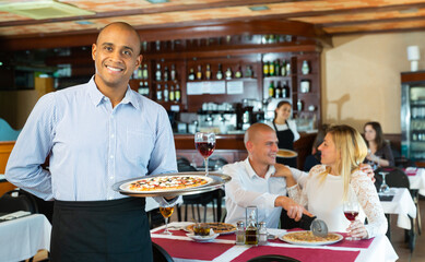 Portrait of friendly Hispanic waiter holding plate with appetizing pizza, warmly welcoming in cozy...