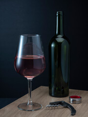 A bottle of red wine and a glass and a corkscrew against a dark background