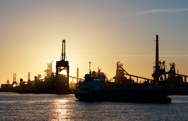 Cranes and smokestacks silhouetted by setting sun behind industrial harbor