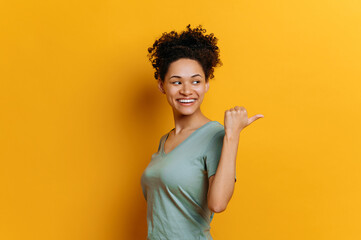 Joyful pretty african american curly haired young woman in basic t-shirt, looks and points finger...