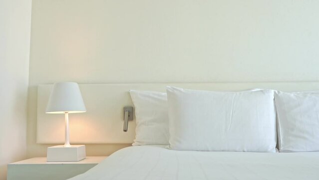 Tilt down on a modern hotel room made in white colors - bed with a set of four pillows and turned on night lap landing on cabinet