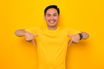 Naklejka premium Smiling young Asian man in casual t-shirt pointing fingers down, offer special discount promo isolated on yellow background. Great offer concept