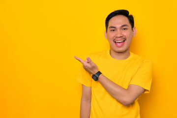 Excited young Asian man in casual t-shirt pointing finger aside at copy space isolated on yellow background. Great offer concept