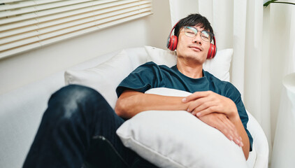 Asian man wearing red glasses and headphones is relaxing on the sofa.