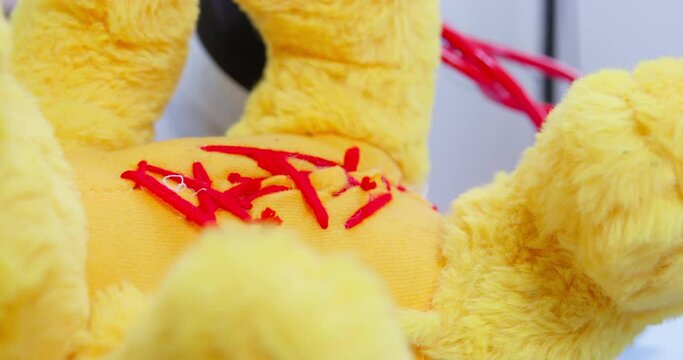 Doctor puts red stitches on torn wound of teddy bear toy on blurred background. Surgeon uses medical scissors to cut thread extreme closeup