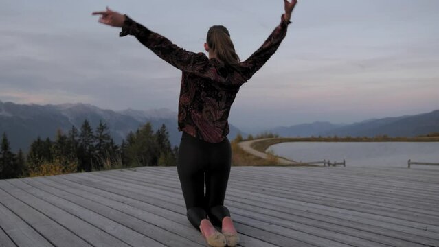 Young woman sitting and dancing on a wooden platform in the mountains at dusk. Alpine mountains, a lake and a cloudy evening sky scenery in the background with fog coming into the frame. Shaky action