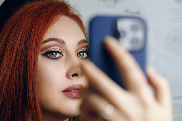 Red-haired woman with bright makeup takes a selfie on the camera of a modern phone. Blogger girl with a smartphone in her hand. Fashionable female hobby.
