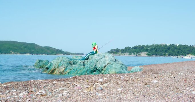 Mermaid with green hair sits on rocks on sea beach covered with pebbles. Mermaid sitting on seashore rocks waves fishtail in sunny weather