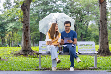 Young Asian and Indian couple enjoy having relaxing time in the rain together in the public park while sitting together on the bench during weekend