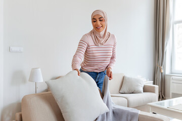 Lovely young Arabic Muslim woman putting soft pillows and plaid on comfy sofa, making her home cozy and warm