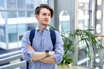 Portrait of happy handsome guy young man student standing indoors at college or university with his hands crossed with backpack and smiling