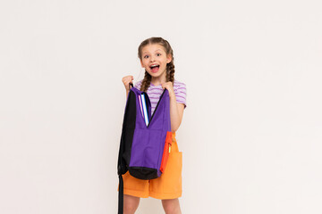 A little girl looks into a school backpack with books. How to pack a backpack for school? A beautiful child with pigtails and shorts on a white isolated background.