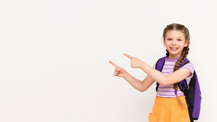 A child with a satchel points with his index finger at your advertisement on a white isolated background. Preparatory summer courses for children. Copy space. - obrazy, fototapety, plakaty