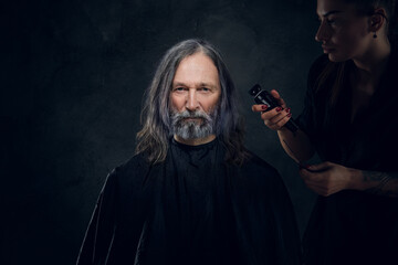 Portrait of long haired elderly man being cutting by female barber against dark background.