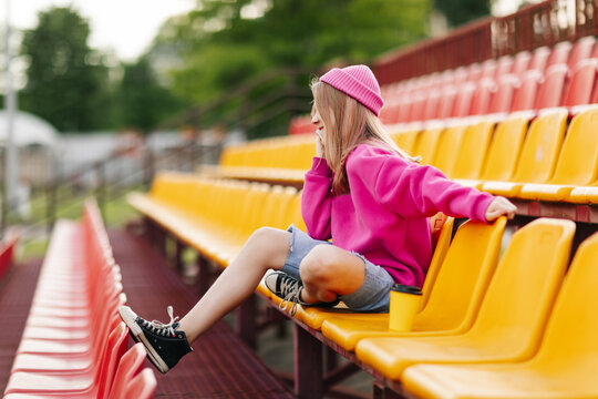 A teenage girl is sitting at the stadium at halftime and talking on the phone with friends. Side view