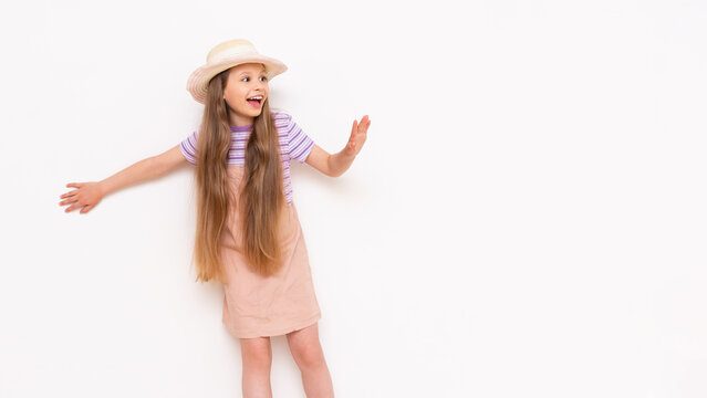 A little girl with long hair in a summer sundress and a straw hat leans against a white walls and looks at your advertisement smiling.Copy space.