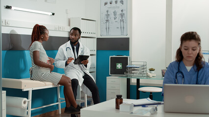Obraz premium Obstetrician using digital tablet to consult patient expecting child at medical appointment. Male doctor taking notes at checkup visit examination, talking to woman with baby bump.