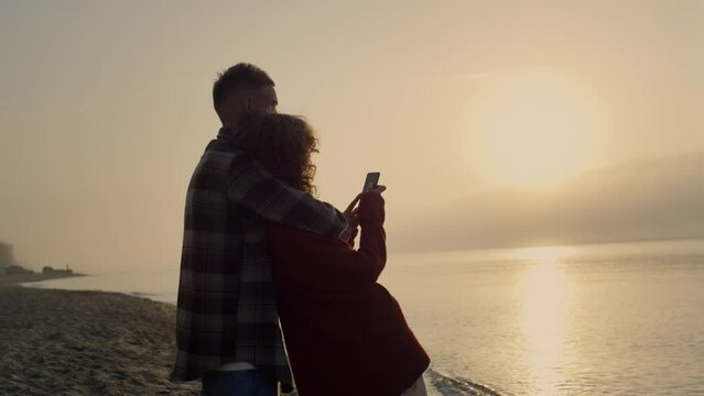 Romantic couple looking sunset on beach. Pretty woman taking photo of ocean