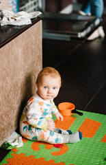 The infant is playing with toys on the rug. Funny baby boy sitting on the floor on kitchen at home. Baby health care and family concept.