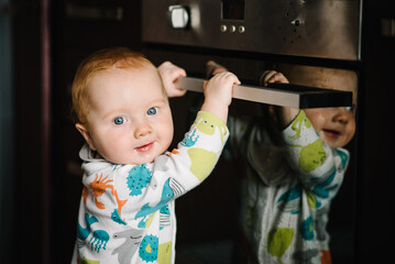 The baby learns to stand near the oven in the kitchen. Little nine months baby boy stands with support at home. Toddler just learnt how to stand on her feet. Close up. © Serhii