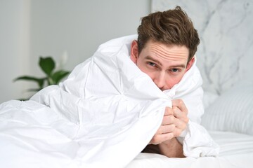 Portrait of handsome sleepy lazy nice man, young guy lying in bedroom in bed covering himself with...