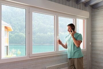 Fototapeta premium Smiled man talking by phone leaning on the window of his apartment.