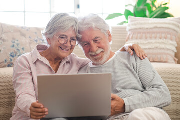 Cheerful caucasian senior couple sitting on the floor at home using laptop, modern retired elderly people surfing the net with computer
