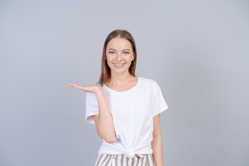 Smiling young woman presenting product holding something on her hand, portrait caucasian beautiful girl in white t-shirt mock up, show showing gesture studio shot on a gray background with copy space