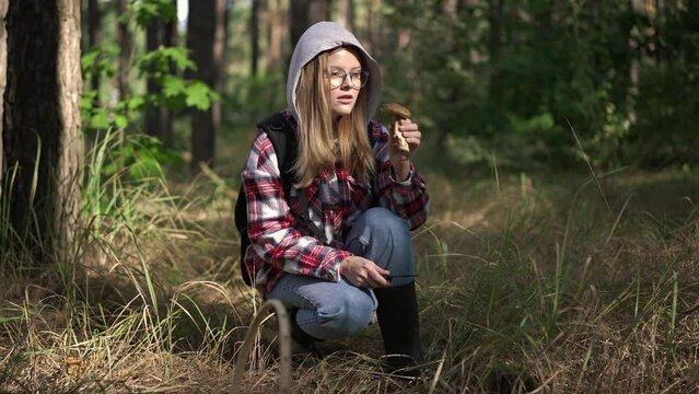 Portrait of young confident woman in eyeglasses cutting mushroom with knife in slow motion sitting in forest outdoors. Happy Caucasian adventurer enjoying mushrooms picking in autumn forest