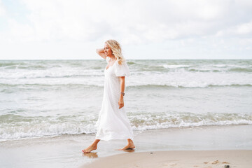 Fototapeta na wymiar Young blonde beautiful woman with long hair in white dress enjoying life on the sea beach