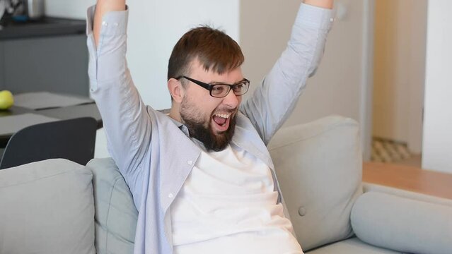 Bearded man in glasses looks emotionally, exclaims his hands, raises his hands and watching interesting TV serial, football game online at home. Concept: home leisure
