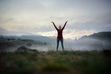 Senior woman doing breathing exercise in nature on early morning with fog and mountains in...